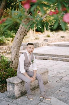 Groom sits on a stone border under a flowering tree in the garden. High quality photo
