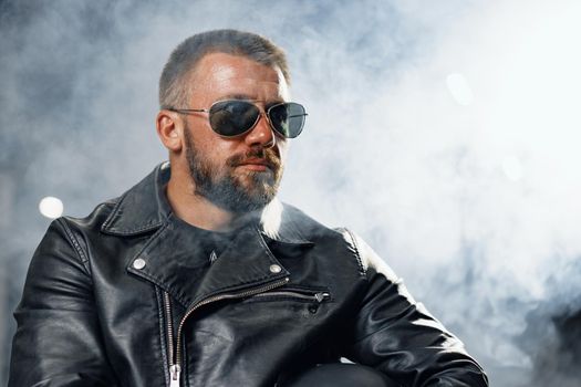 Portrait of bearded man motocyclist in dark sunglasses on dark building background