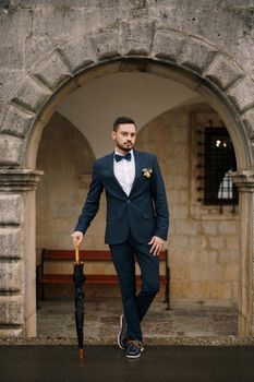 Groom stands under the arch of an old building leaning on an umbrella cane. High quality photo