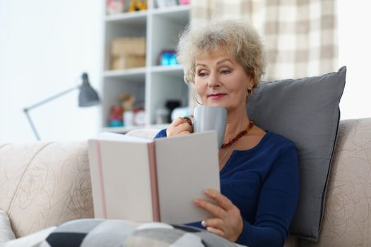 Portrait of old lady reading book and drinking cup of coffee being comfy on sofa at home. Attractive senior woman relaxing on weekend. Spare time concept