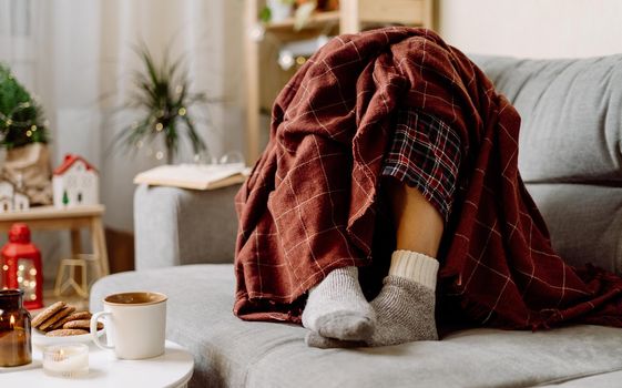 Cozy woman legs in knitted winter warm socks under checkered plaid drinking hot cocoa or coffee in mug, during resting on couch at home. Christmas holidays with candle, decor, book and cookies