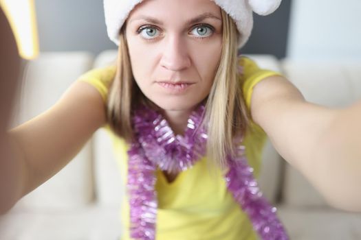 Portrait of beautiful serious blonde young woman posing on camera in santa claus hat. Celebrating new year alone at home. Holiday, christmas mood concept
