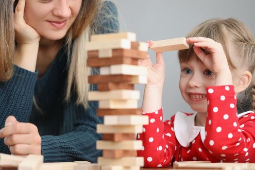 Portrait of pretty little kid pulling wooden block out of tower and smile. Mother join daughter in funny board game at home. Parenthood, childhood concept