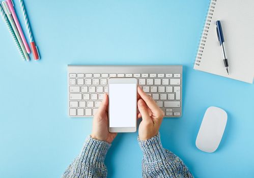 Mobile phone with blank white screen display in hand. Bright blue modern desk with smartphone. Top view. Copy space, mock up.