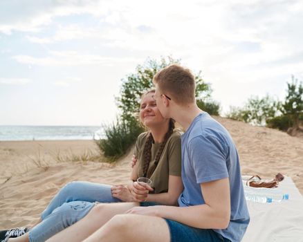 Picnic on beach with food and drinks. Young boy kissing girl sitting on sand. Teens couple. Sunny summer day, rest.