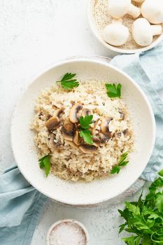 Risotto with mushrooms in plate. Rice porridge with mushrooms and parsley. White table, spoons, mushrooms. Hot dish, italian cuisine, top view, vertical