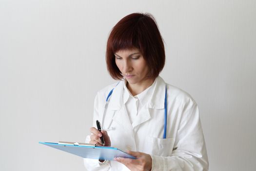 Female adult doctor stands on white background. Beautiful woman, thinking, writing. Clipboard in his hand, stethoscope around his neck .