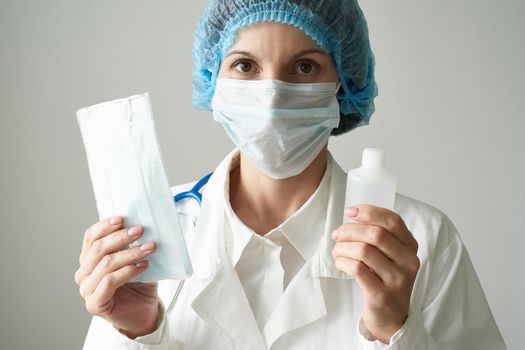 Female doctor in medical mask and uniform offers mask and disinfectant in bottle to prevent from coronavirus. Covid-19 protection, medical information