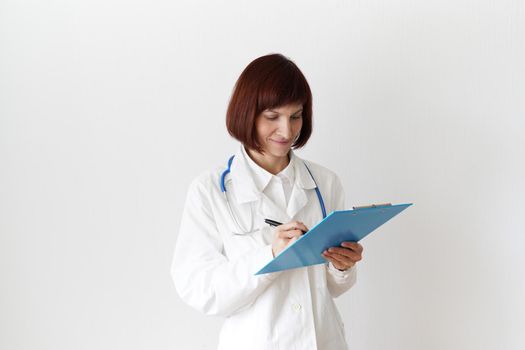 Female adult doctor stands on white background. Beautiful woman, thinking, writing. Clipboard in his hand, stethoscope around his neck .