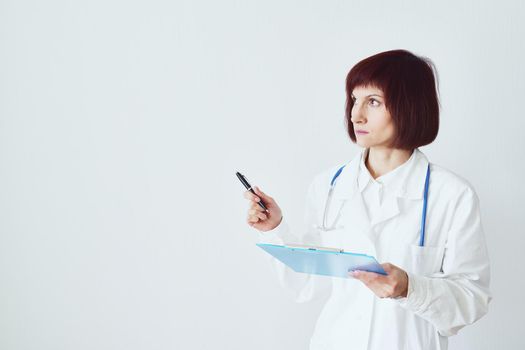 Female adult doctor stands on white background shows, makes a hand gesture. Beautiful woman, thinking. Clipboard in his hand, stethoscope around his neck .