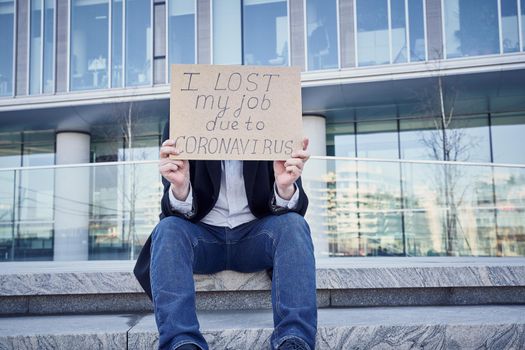 Job loss due to COVID-19 virus pandemic concept. Unrecognizable man holds sign I lost my job because of coronavirus. Male sitting on stairs of building, former place of work in business center