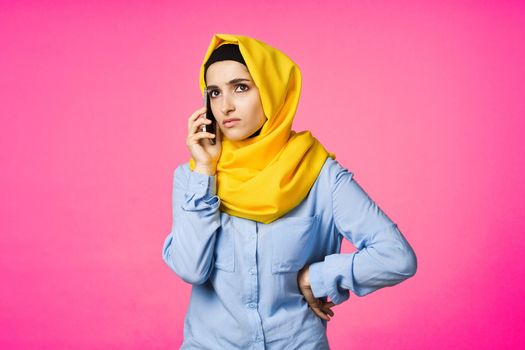 muslim woman in yellow hijab talking on the phone technology pink background. High quality photo