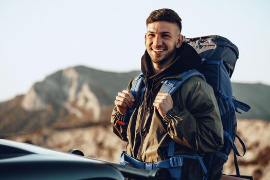 Portrait of a young traveler man in hiking equipment standing near his off-road car at dawn