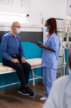 Physician woman nurse with medical protective face mask against coronavirus discussing sickness symptoms with retired senior man typing disease diagnosis on tablet in hospital office. Medicine concept