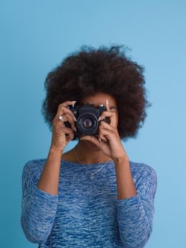 portrait of a smiling pretty african american girl taking photo on a retro camera isolated over blue background