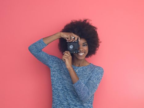 portrait of a smiling pretty african american girl taking photo on a retro camera isolated over pink background