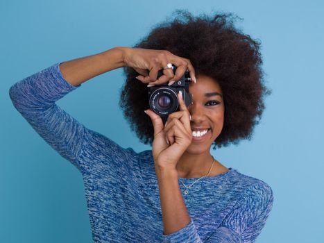portrait of a smiling pretty african american girl taking photo on a retro camera isolated over blue background