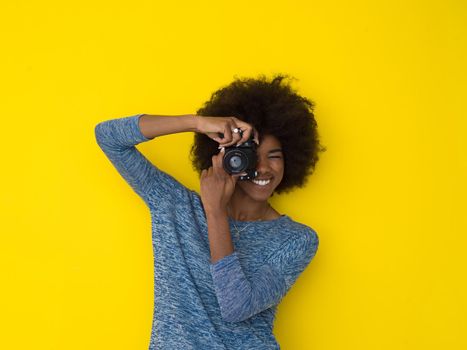 portrait of a smiling pretty african american girl taking photo on a retro camera isolated over yellow background