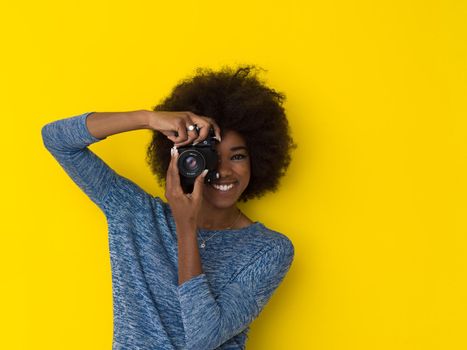 portrait of a smiling pretty african american girl taking photo on a retro camera isolated over yellow background