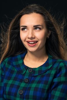 Portrait of cheerful attractive modern woman with long hair, expressing positive emotions while standing over black background. Young woman in blue checkered dress