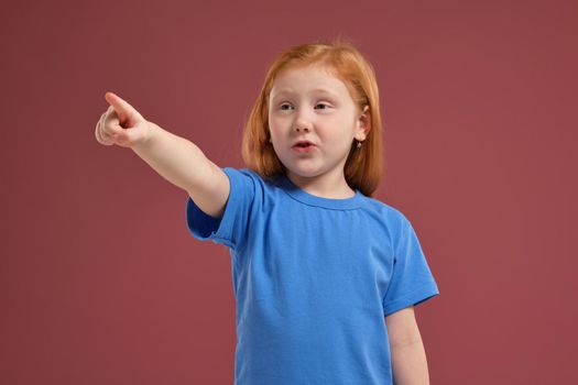 Portrait of cute redhead emotional little girl in blue t-shirt on red background, pointing on something with her finger