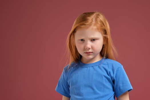 Portrait of cute redhead emotional little girl in blue t-shirt on red background. She looks very upset and about to cry
