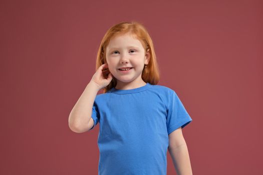 Portrait of cute redhead emotional little girl in blue t-shirt on red background. Beautiful studio portrait of female little caucasian person. SHe looks very happy