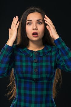 Portrait of cheerful attractive modern woman with long hair, looking at camera and expressing surprise emotions while standing over black background. Young woman in blue checkered dress