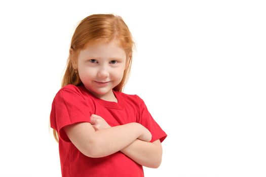 Portrait of cute redhead emotional little girl in red t-shirt isolated on a white. Beautiful studio portrait of female little caucasian person with crossed hands looking on camera