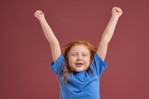 Portrait of cute redhead emotional little girl in blue t-shirt on red background. She is tired and wants to sleep, yawning and stretching