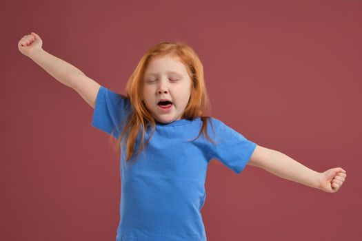 Portrait of cute redhead emotional little girl in blue t-shirt on red background. She is tired and wants to sleep, yawning and stretching