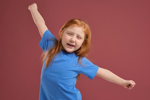 Portrait of cute redhead emotional little girl in blue t-shirt on red background. She is tired and wants to sleep, yawning and stretching