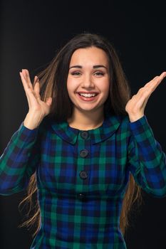 Portrait of cheerful attractive modern woman with long hair, looking at camera and expressing positive emotions while standing over black background. Young woman in blue checkered dress