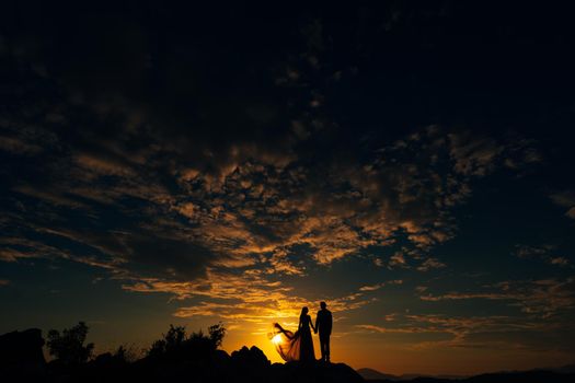 Groom and bride stand on the mountain against the background of the sunset sky. High quality photo