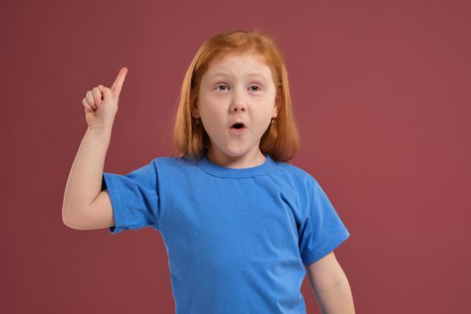 Portrait of cute redhead emotional little girl in blue t-shirt on red background. I have an idea! Eureka bingo