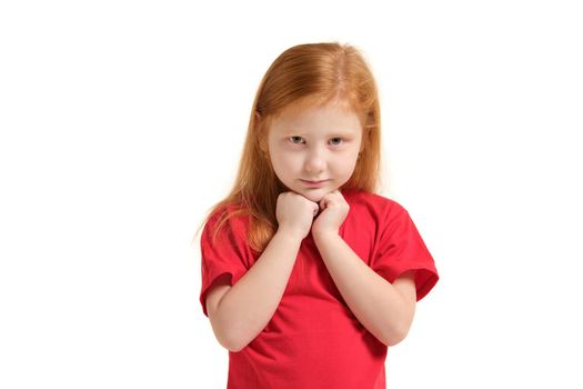 Portrait of cheerful caucasian little girl with red hair posing in studio with looking to the camera. Girl dressed in red T-shirt keeping palms pressed together, having begging look.