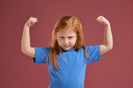 Portrait of cute redhead emotional little girl in blue t-shirt on red background. She frowns and try to look scare