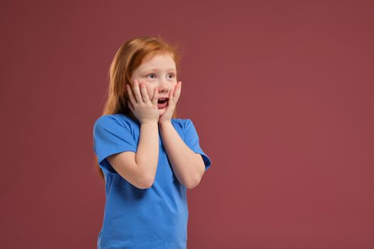 Portrait of cute redhead emotional little girl in blue t-shirt on red background. Beautiful studio portrait of female little caucasian person holding her head stressed
