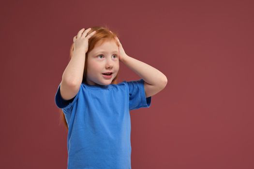 Portrait of cute redhead emotional little girl in blue t-shirt on red background. Beautiful studio portrait of female little caucasian person holding her head stressed