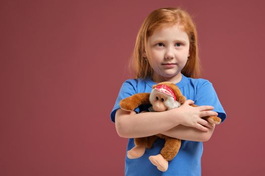 Portrait of cute redhead emotional little girl in blue t-shirt on red background. She hugs toy plush monkey