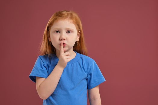 Redhead girl in blue t-shirt putting finger up to lips saying shahs on red background
