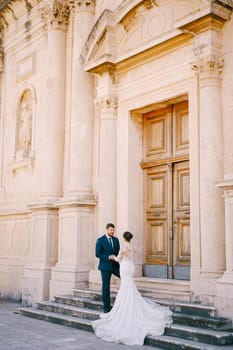 Bride and groom stand on the stone steps in front of the wooden door of the ancient church. High quality photo