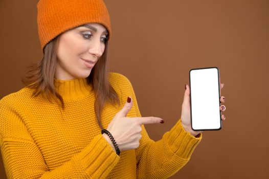 Portrait of an attractive caucasian young woman in a yellow sweater and hat holding a smartphone. in her hands and points her finger at the cut out phone display.