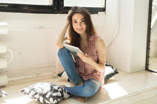 Technology and people concept - Young beautiful woman sitting on the wooden sill with tablet and pillows.