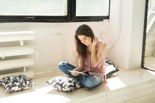 Technology and people concept - young woman sitting on the wooden sill with tablet and pillows.