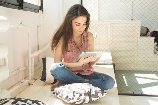 Technology and people concept - Young beautiful woman sitting on the wooden sill with tablet and pillows.