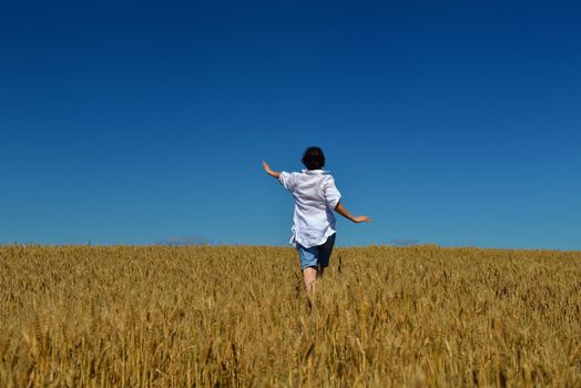Young woman standing jumping and running  on a wheat field with blue sky in  background at summer day representing healthy life and agriculture concept