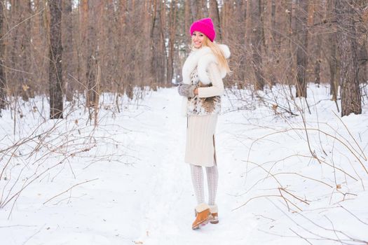 Fashion, season and people concept - Happy blonde woman in a pink hat and gloves and sweater in the winter forest is smiling.