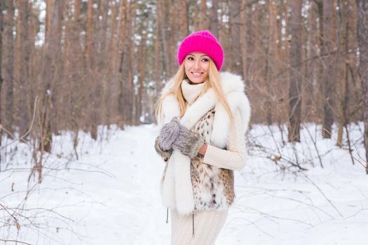 Winter and people concept - Happy blonde woman in a pink hat and gloves and sweater in the winter forest is smiling and looking aside.