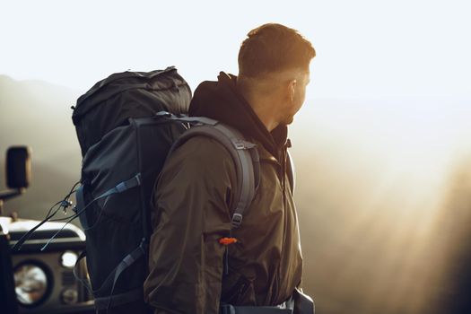 Portrait of a young traveler man in hiking equipment standing near his off-road car at dawn